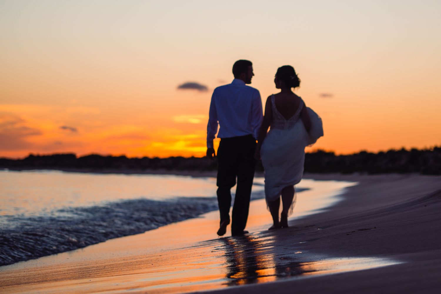 Couple walking hand in hand on a beach at sunset, silhouetted against a vibrant orange and red sky.
