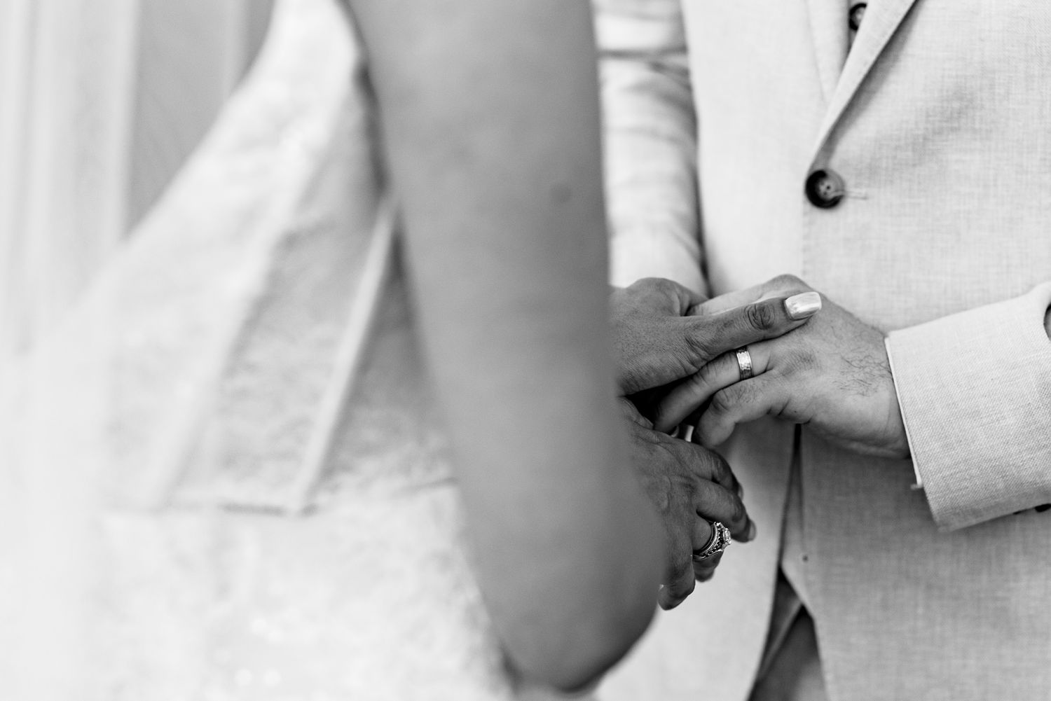 Black and white photo of bride and groom holding hands, wedding ring visible on groom's finger.