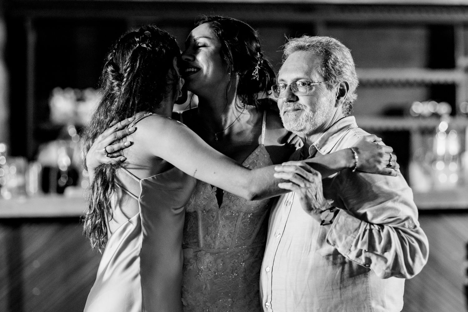 Black and white photo of two women embracing an older man during a wedding reception dance.