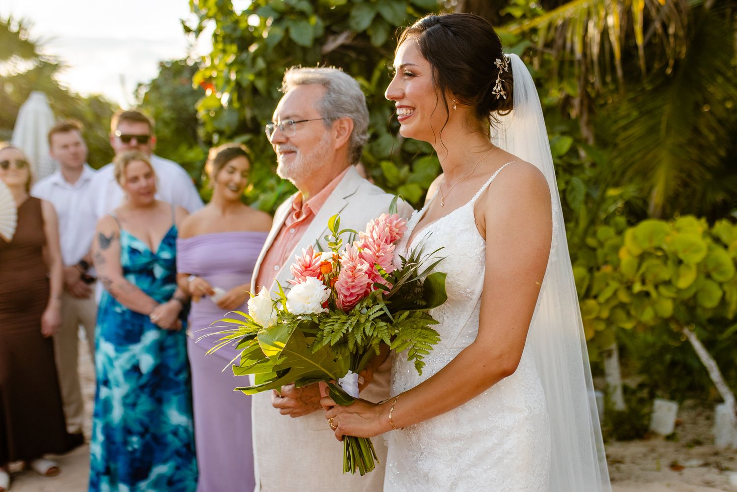 Bride in white gown holding tropical bouquet stands with older man at outdoor beach wedding ceremony.
