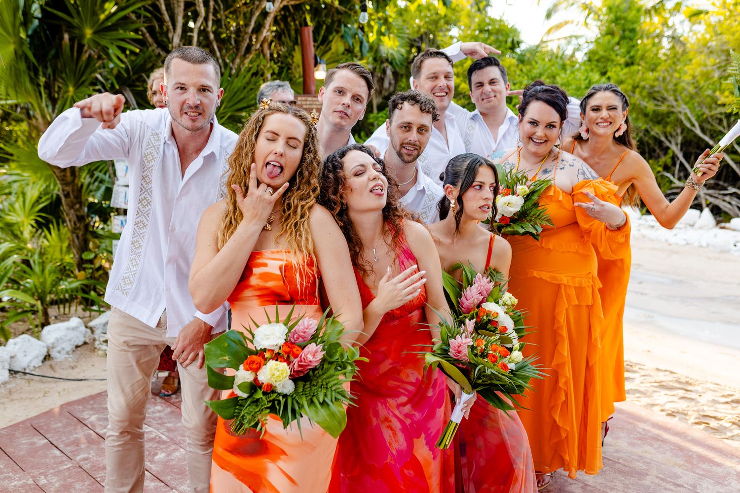 Joyful wedding party in colorful orange and pink attire posing playfully outdoors with tropical bouquets.