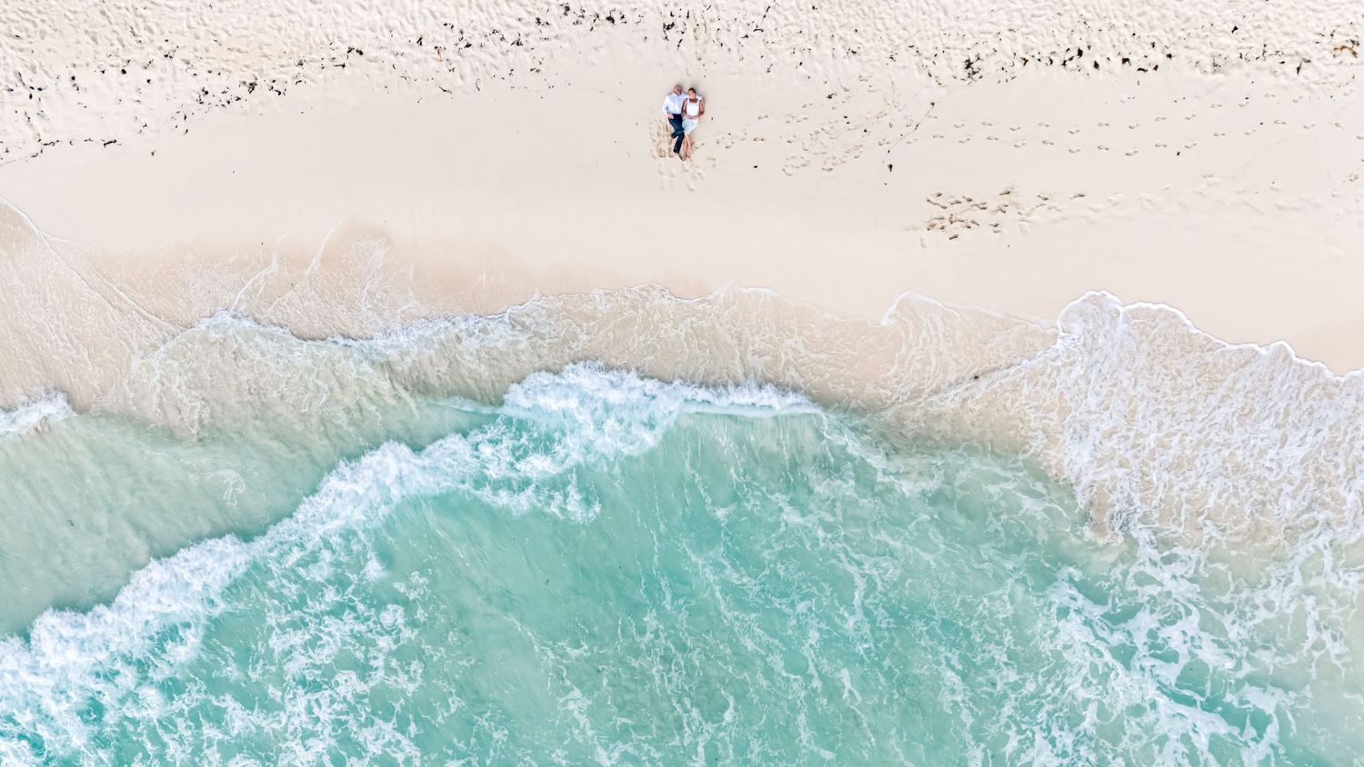Aerial view of turquoise ocean waves crashing onto a sandy beach with white foam patterns visible from above.