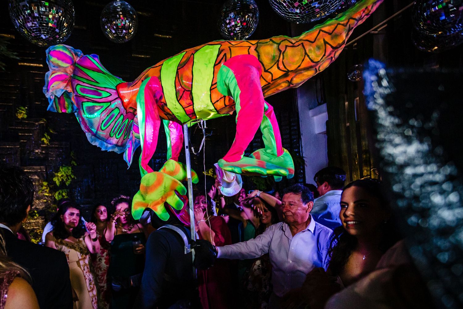 Colorful neon dinosaur balloon float at a lively party with disco balls and dancing guests in dark venue.