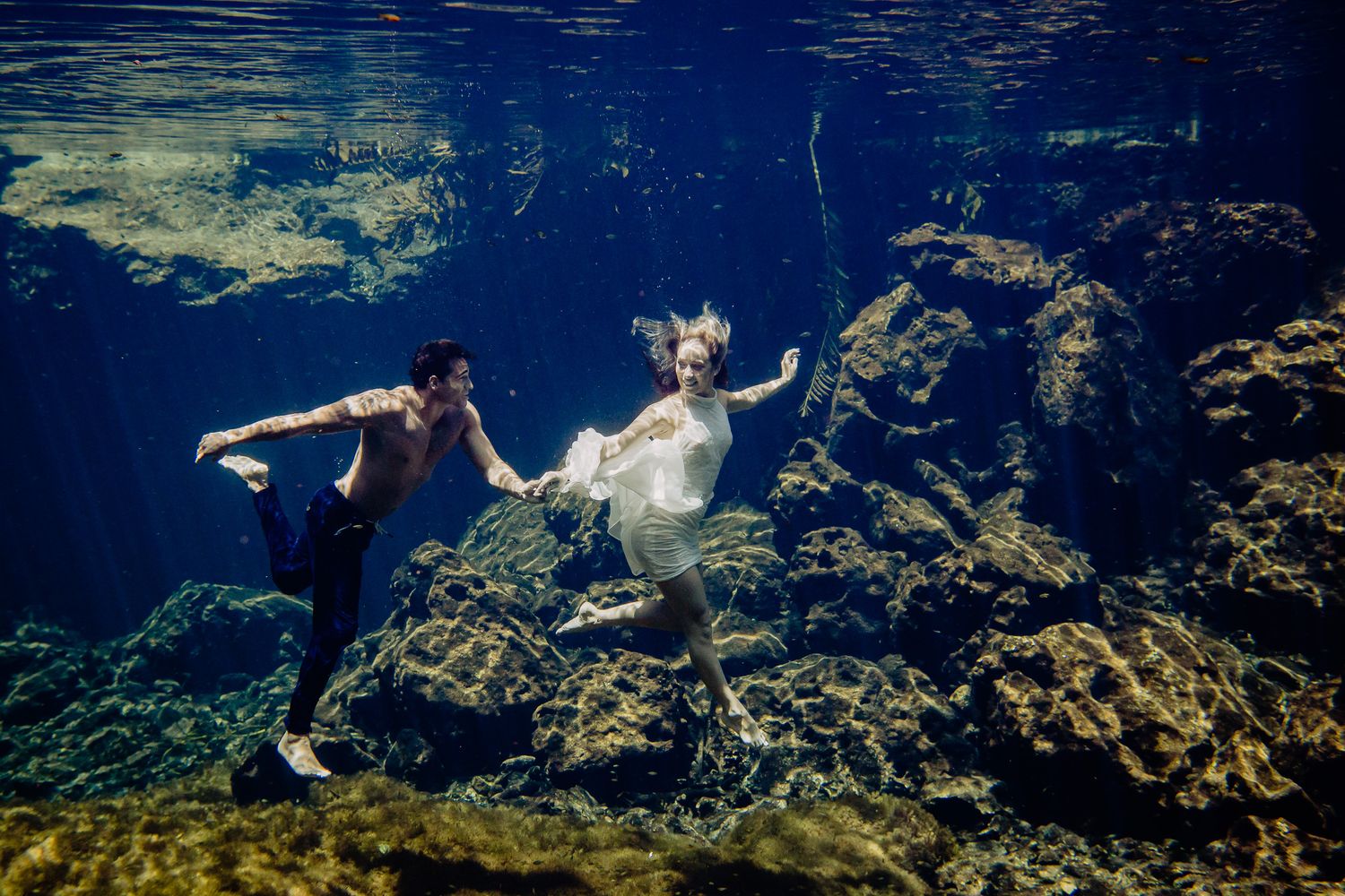 Couple in wedding attire swimming underwater among rocks and coral in a stunning underwater engagement photo.
