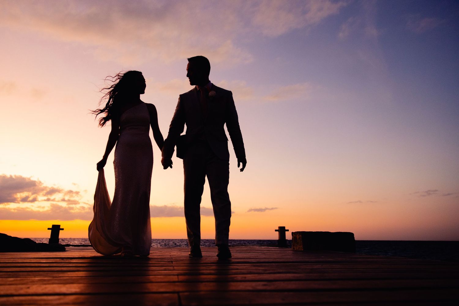Silhouettes of a couple holding hands walking on the beach at sunset against an orange and purple sky.