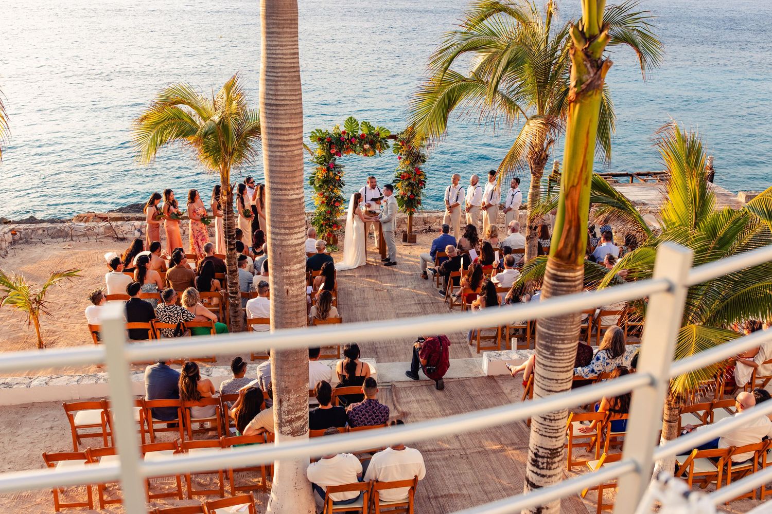 Aerial view of outdoor beach wedding ceremony with guests seated near ocean at sunset.