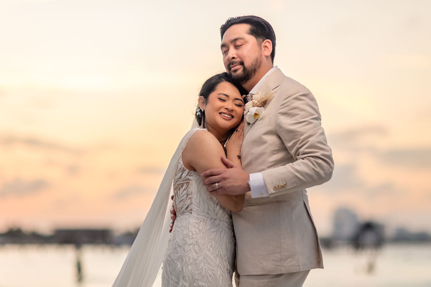 Bride and groom embracing tenderly at sunset by the water, groom in beige suit, bride in lace gown.