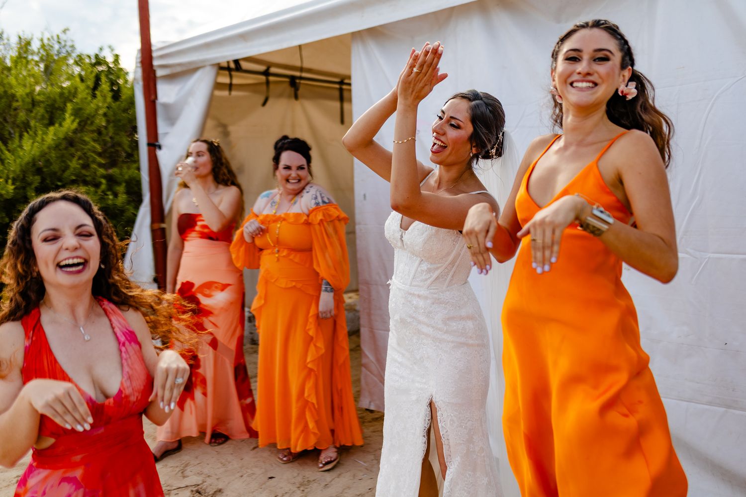 Joyful bridesmaids in vibrant orange and red dresses dancing and laughing together at an outdoor wedding celebration.