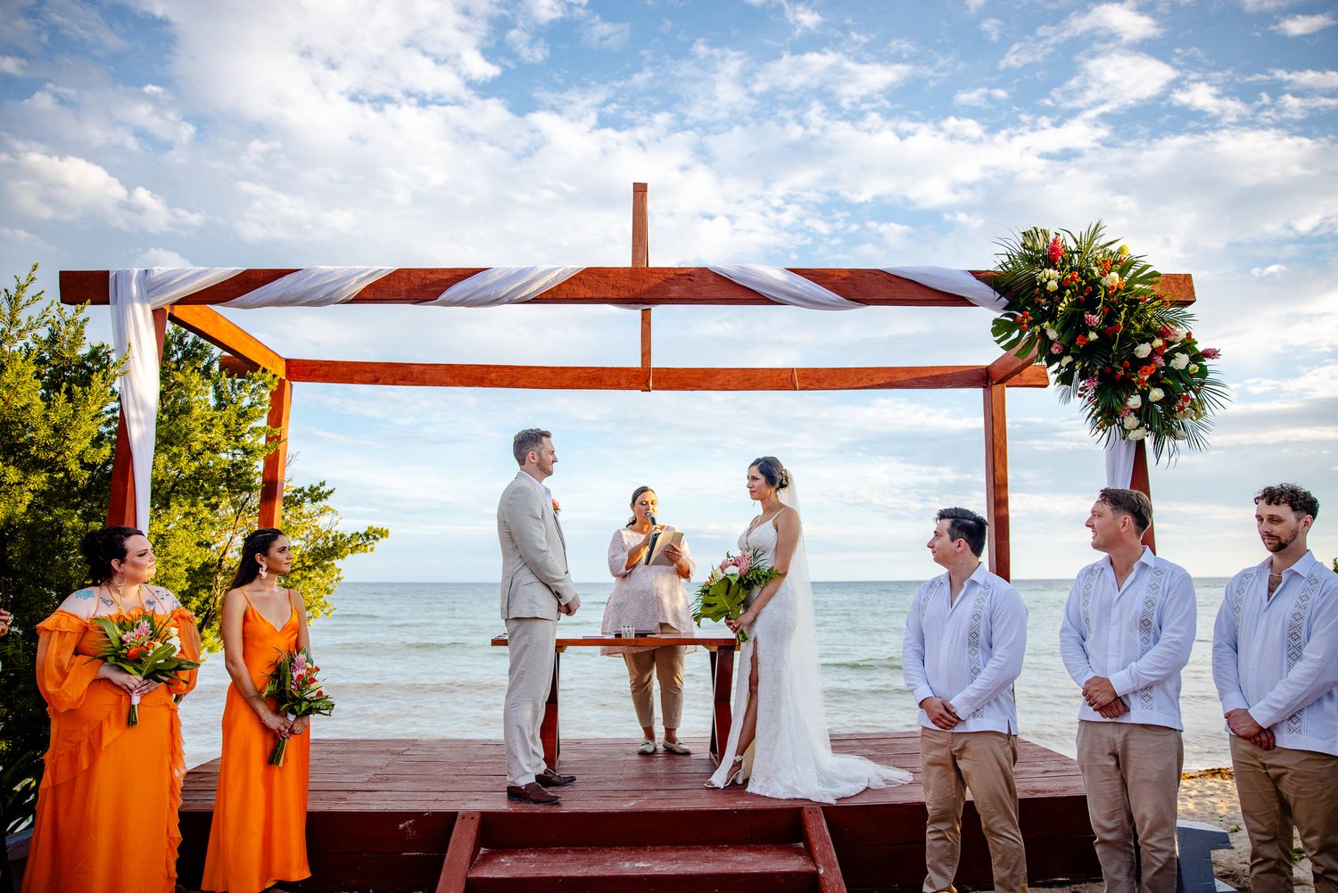 Beachside wedding ceremony under orange canopy with couple, officiant, bridesmaids in orange dresses, and groomsmen by the sea.