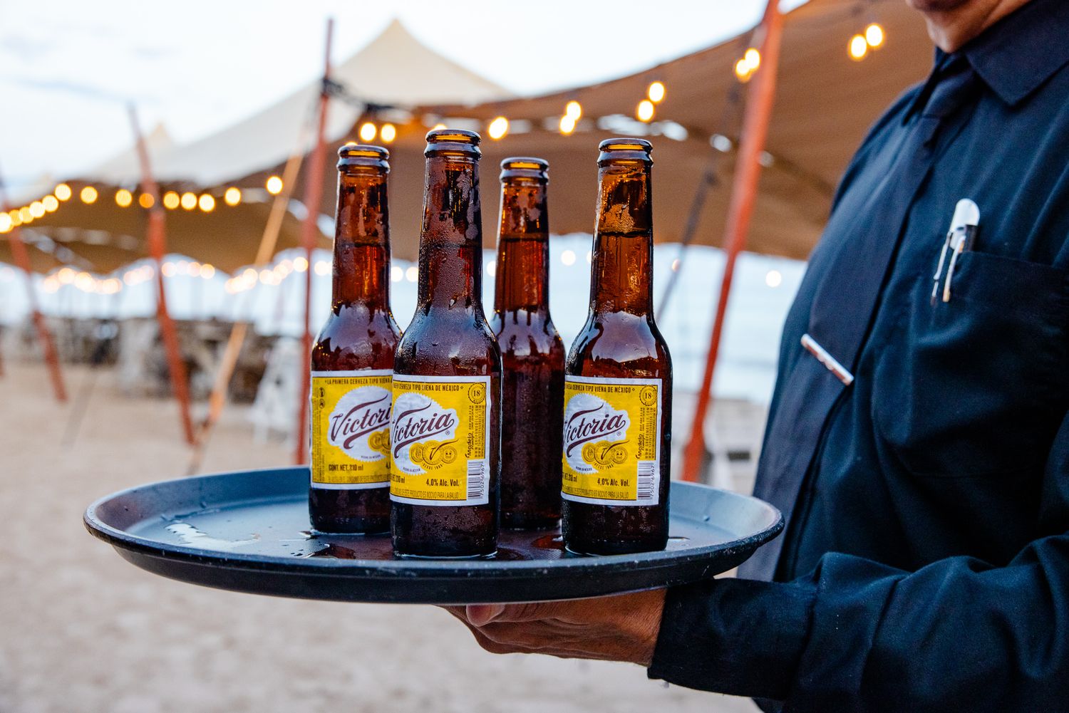 Waiter serving three cold Corona beer bottles on a tray at an outdoor beach event under string lights.