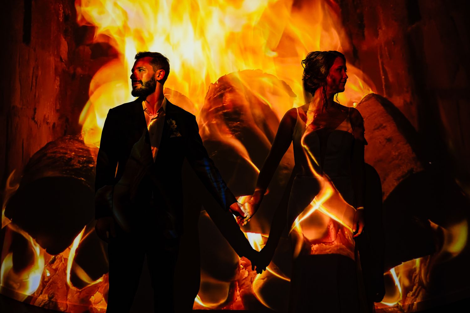 Couple silhouetted against dramatic flames, holding hands in dark formal attire, creating a striking fire backdrop.