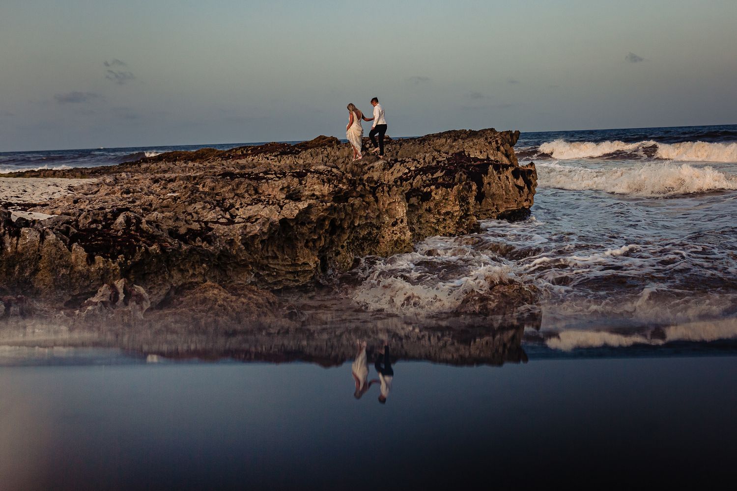 Couple standing on rocky shore with ocean waves, their reflection visible in tide pools at sunset.