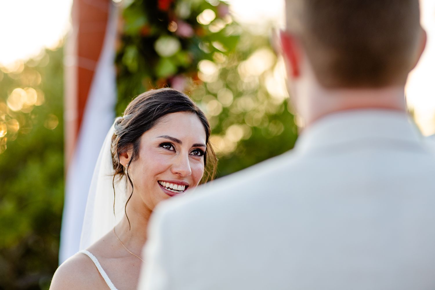 Smiling bride gazes lovingly at groom during outdoor wedding ceremony with golden sunset light.