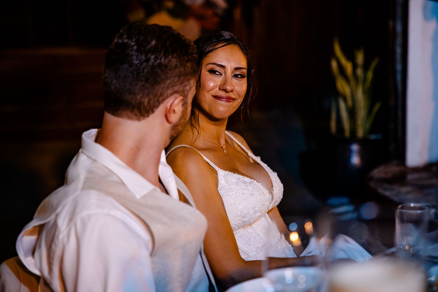 Bride in white lace dress smiling warmly at groom during wedding reception dinner by candlelight.