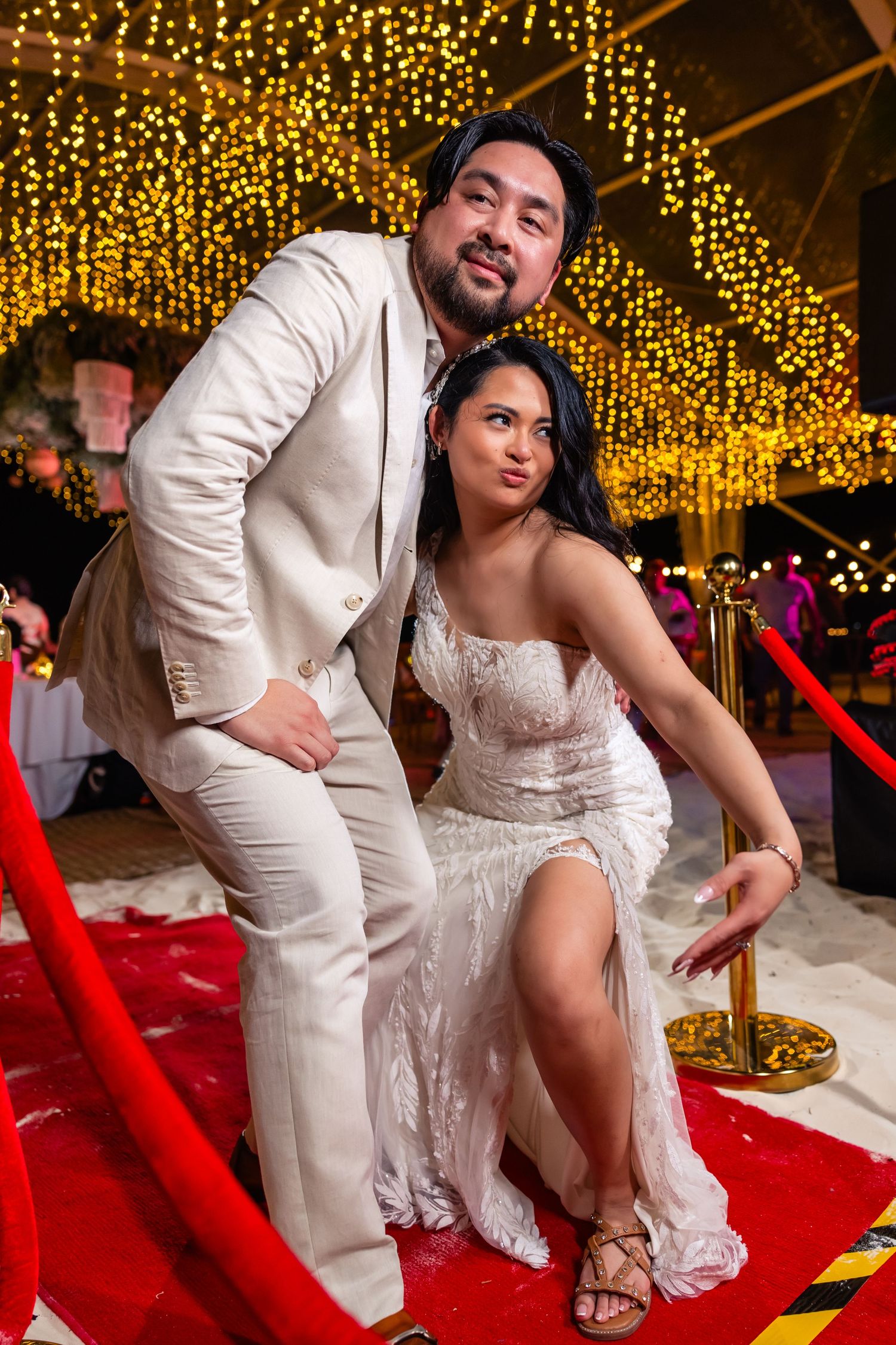 Playful bride and groom posing on red carpet under golden fairy lights at festive wedding reception.