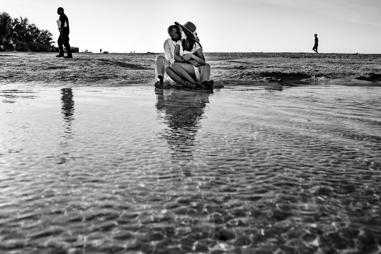 Black and white photograph of people sitting in shallow beach water with reflections and silhouettes in the background.