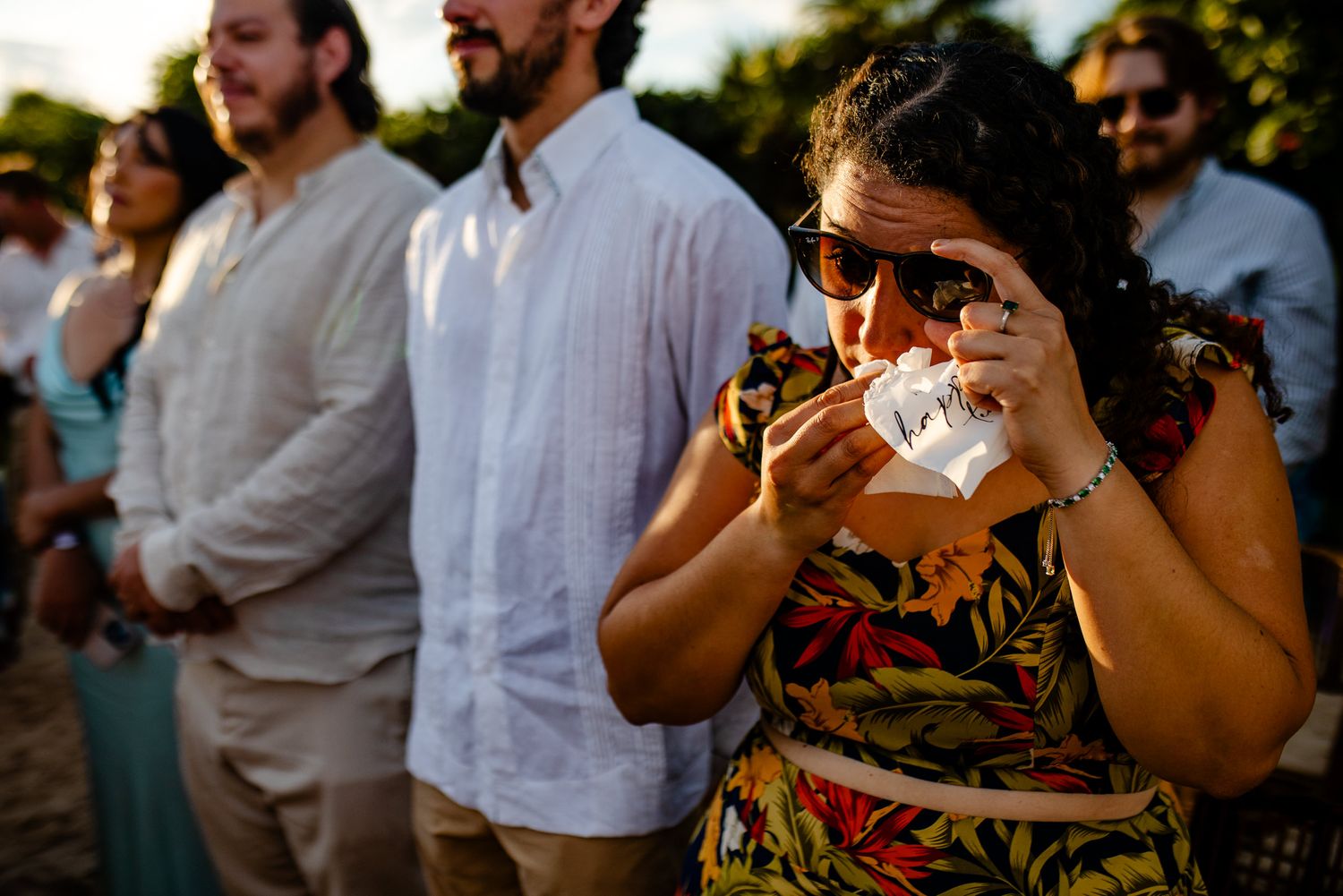 Emotional wedding guest wiping tears with tissue outdoors at sunset ceremony, wearing floral dress and sunglasses.