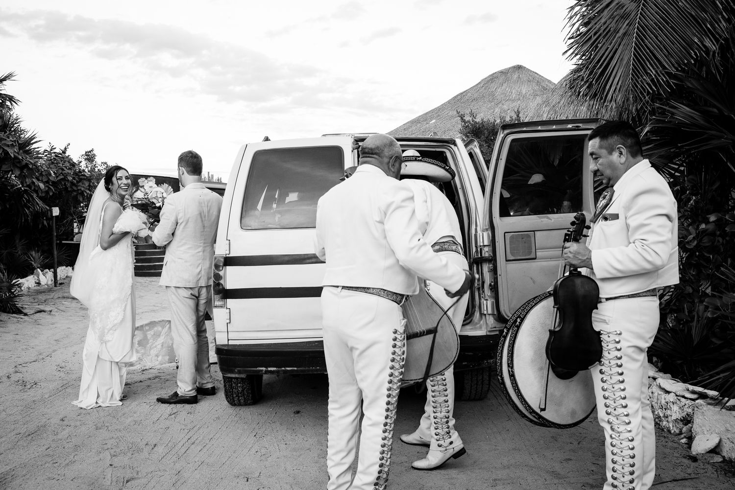 Black and white photo of newlyweds with mariachi band loading van at Mexican beach wedding venue.