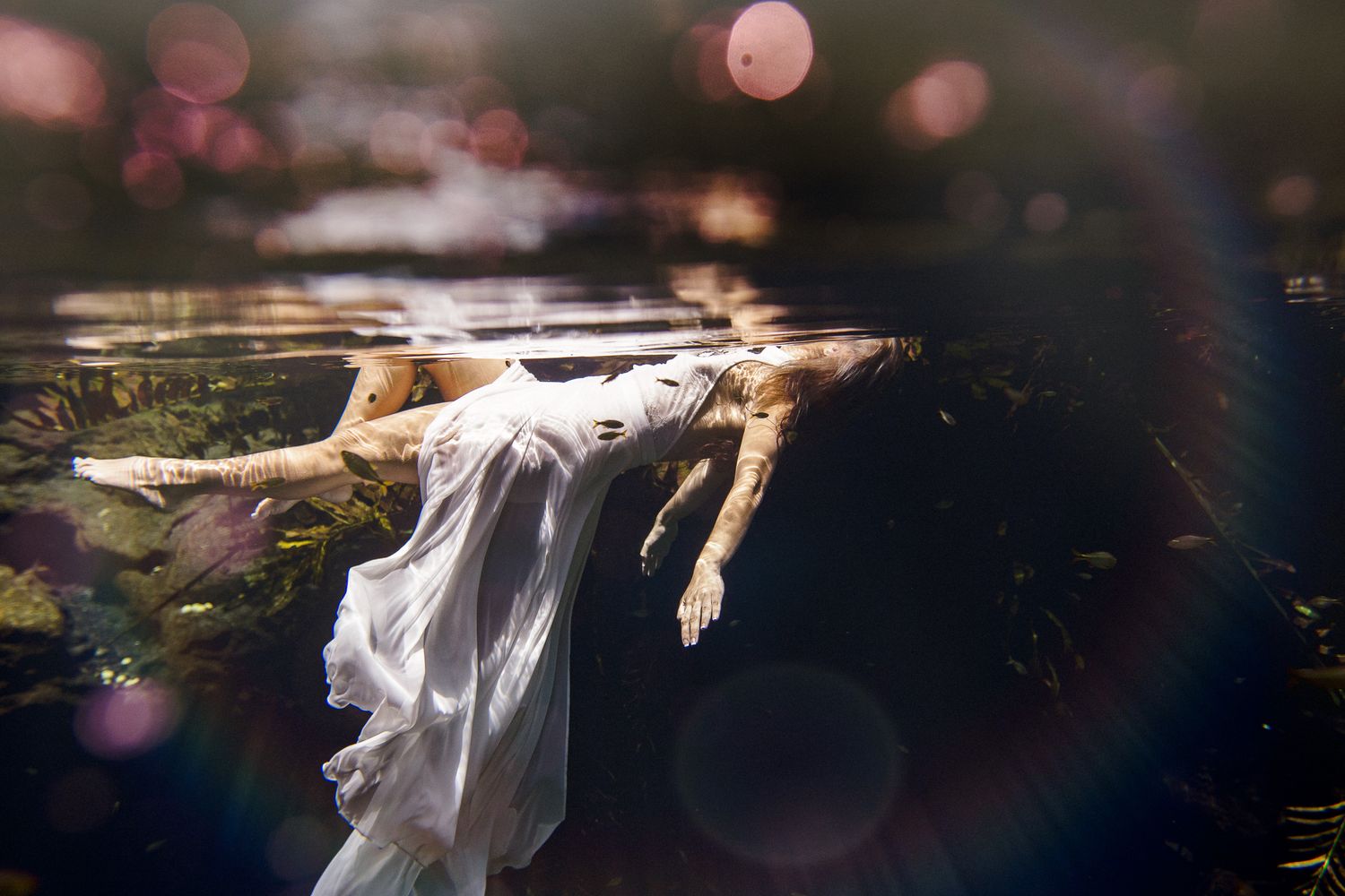 Person in white dress floating underwater with bokeh light effects creating a dreamy, ethereal atmosphere.