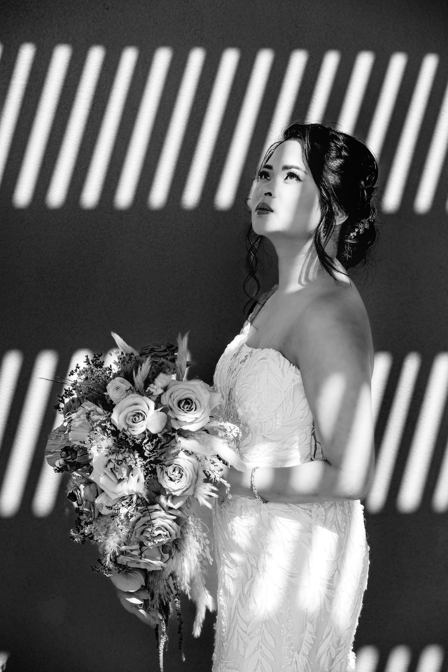 Black and white portrait of a bride in strapless lace gown holding a bouquet with dramatic striped light shadows.