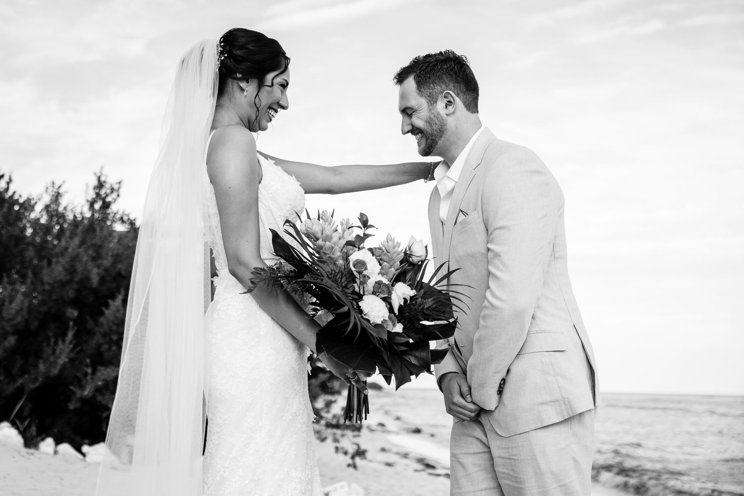 Black and white photo of a bride and groom sharing a joyful moment on a beach, bride holding a tropical bouquet.