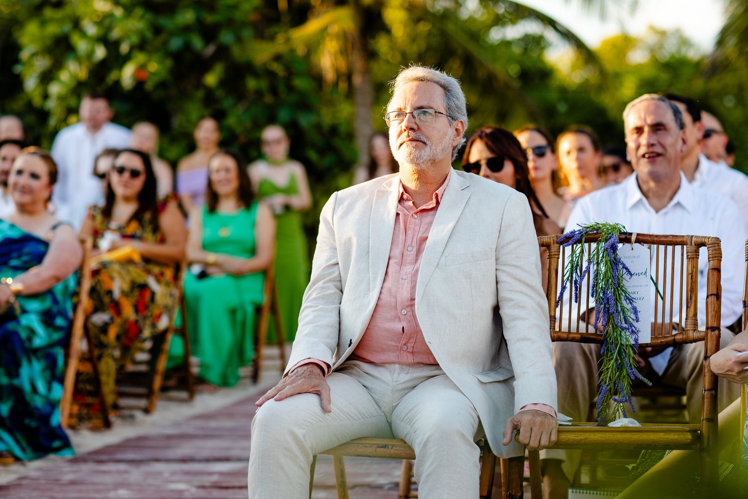 Man in beige suit seated at outdoor wedding ceremony with guests in background.