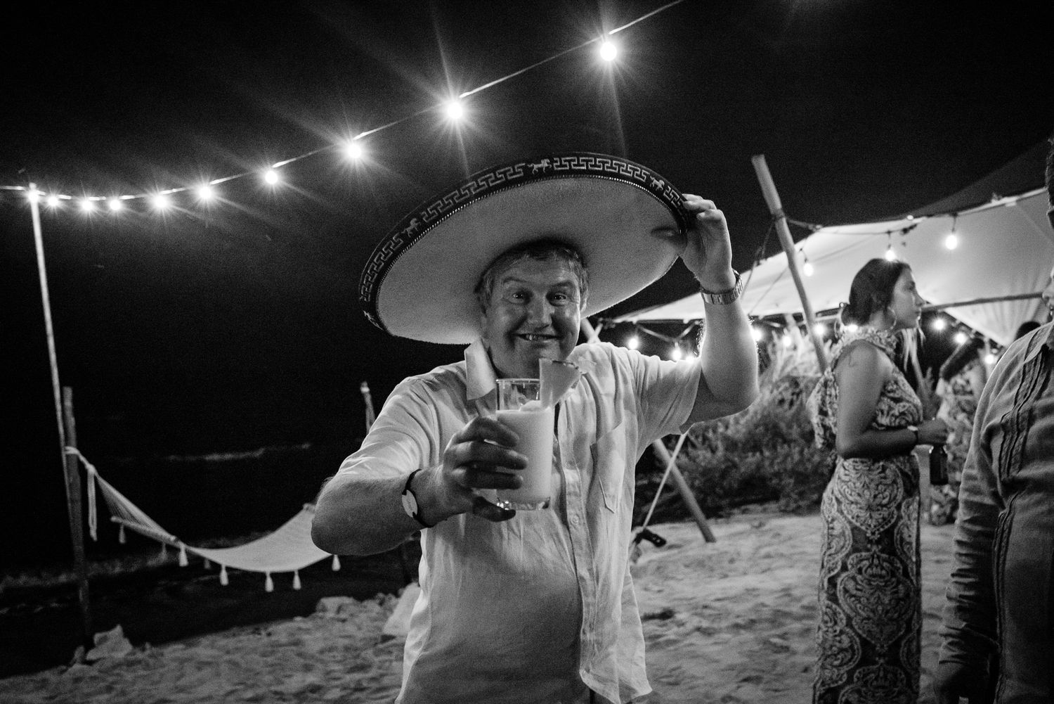 Man in sombrero holding drink at beach wedding party with string lights at night.
