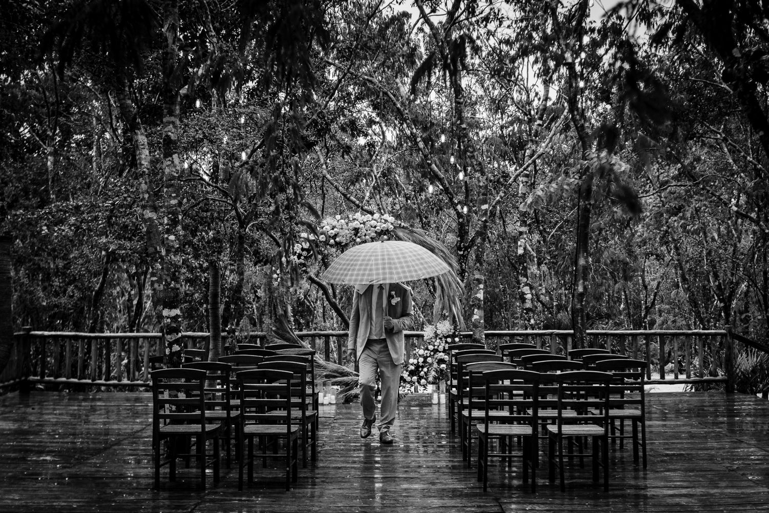 Black and white photo of a person holding an umbrella in the rain at an outdoor wedding venue with empty chairs.