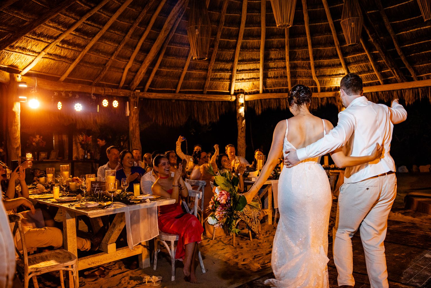 Bride and groom share first dance under thatched roof at tropical beach wedding reception with guests celebrating.
