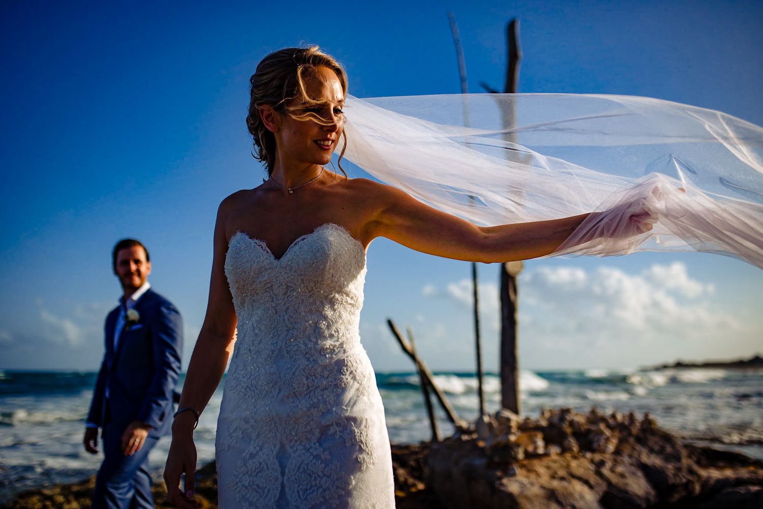Bride in lace gown with flowing veil by ocean, groom in blue suit behind her on rocky shore under blue sky.