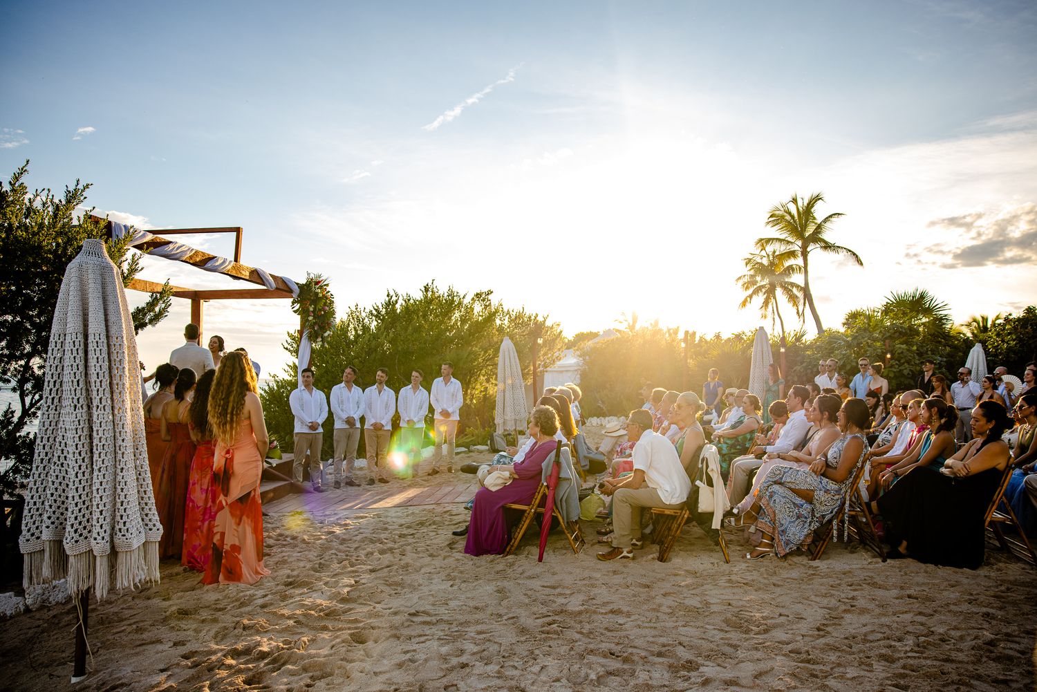 Outdoor beach wedding ceremony at sunset with guests seated on sand, tropical palm trees, and golden sunlight in the background.