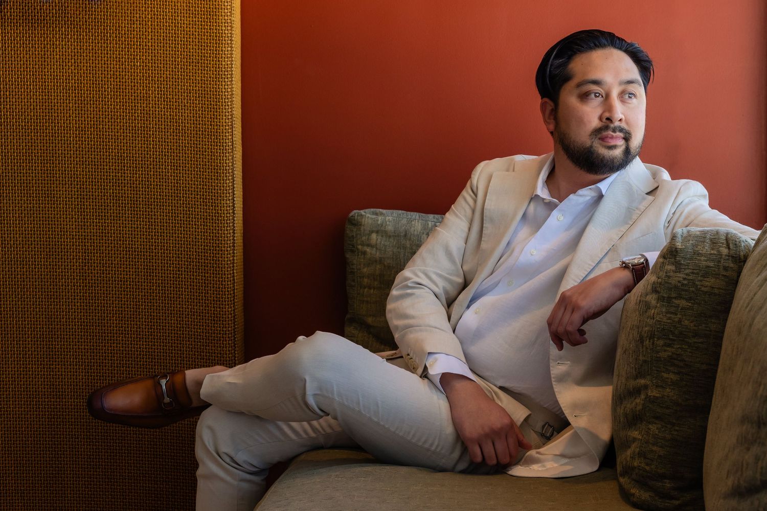 Stylish man in beige blazer sitting relaxed on green sofa against orange wall in modern interior setting.
