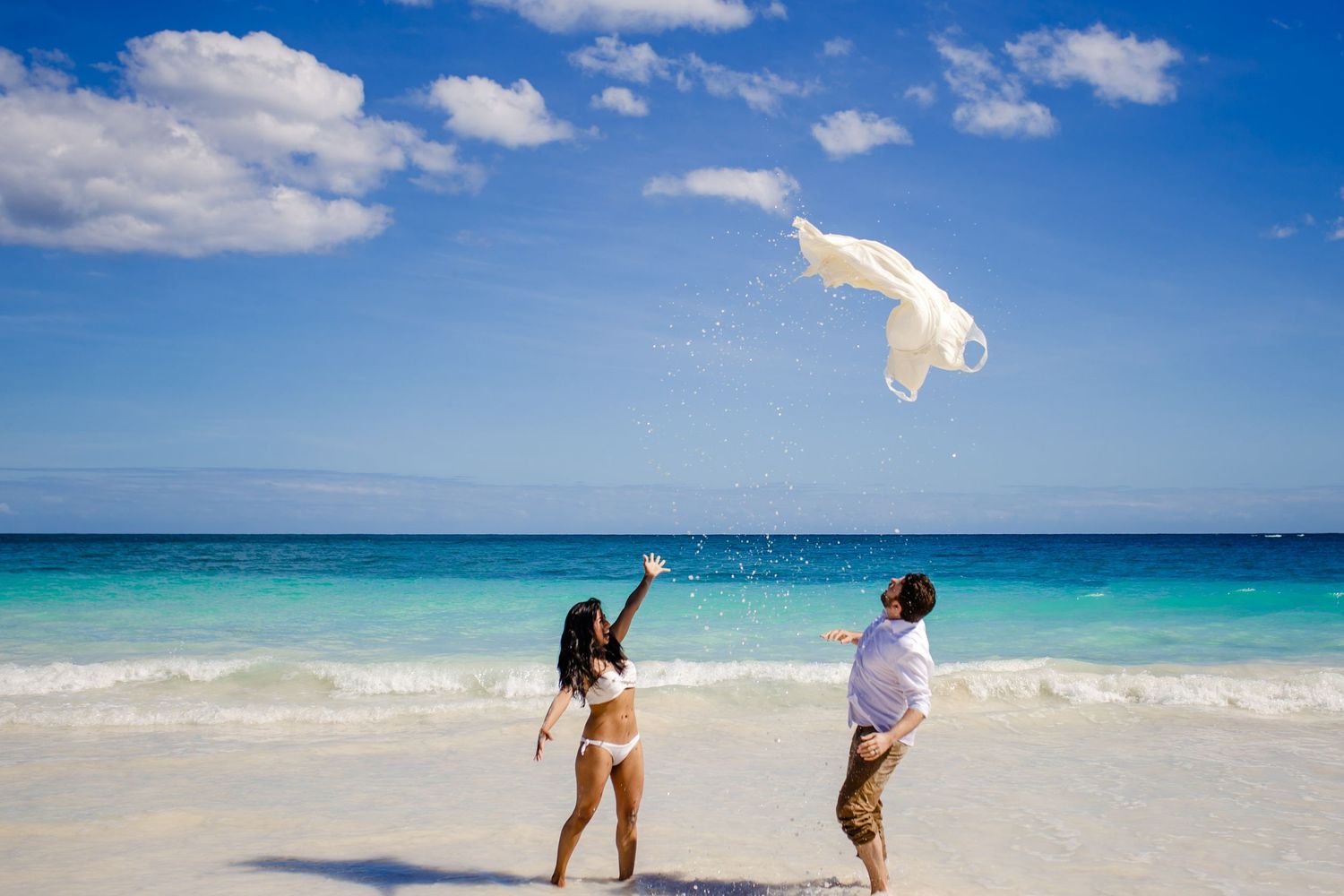 Couple tossing a white veil on a tropical beach with turquoise water and blue sky.