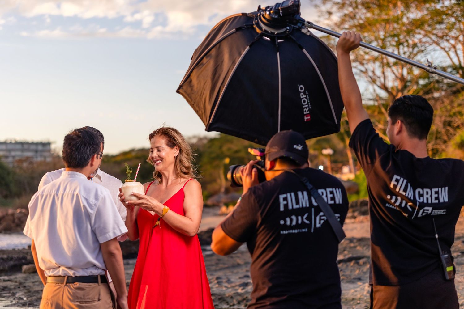 Film crew photographing a woman in a red dress outdoors during golden hour with a softbox light.