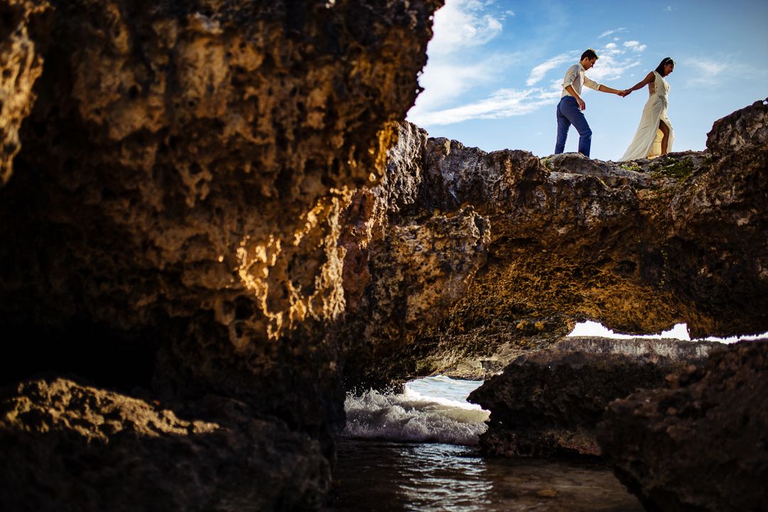 Bride and groom hold hands on rocky coastal cliffs with ocean waves below during dramatic outdoor wedding photo session.