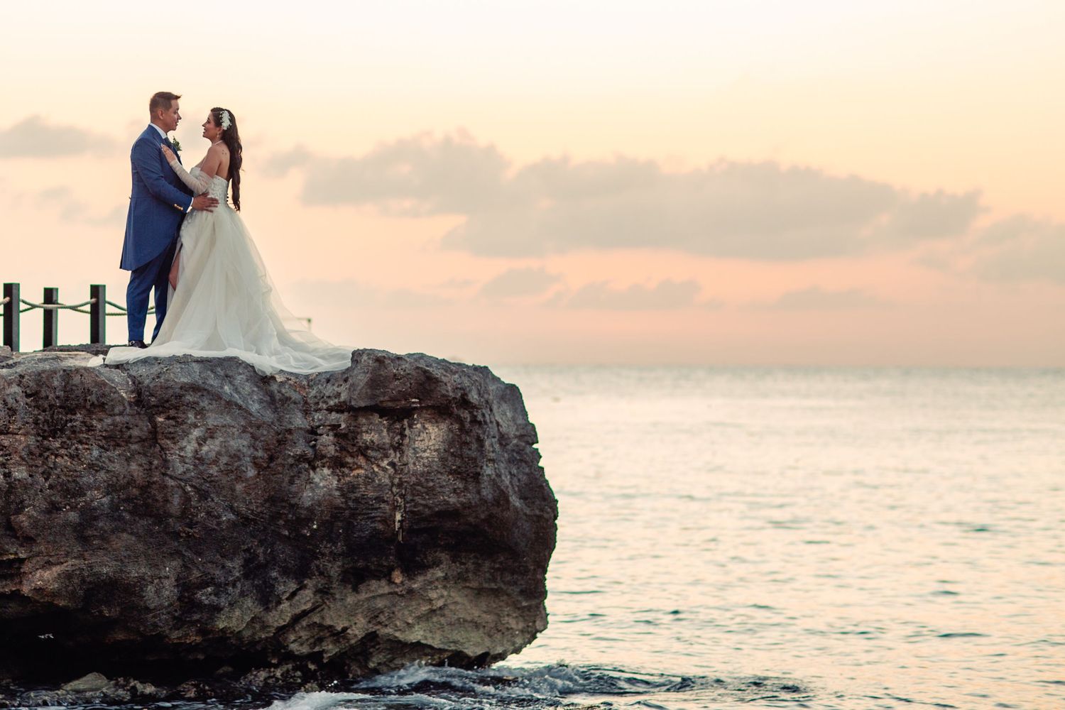 A couple in wedding attire stands on a rocky cliff overlooking the ocean during a dreamy sunset.