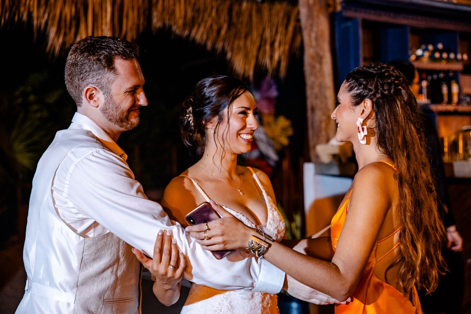 Bride and groom share a joyful moment with a guest in orange dress at tropical wedding reception.