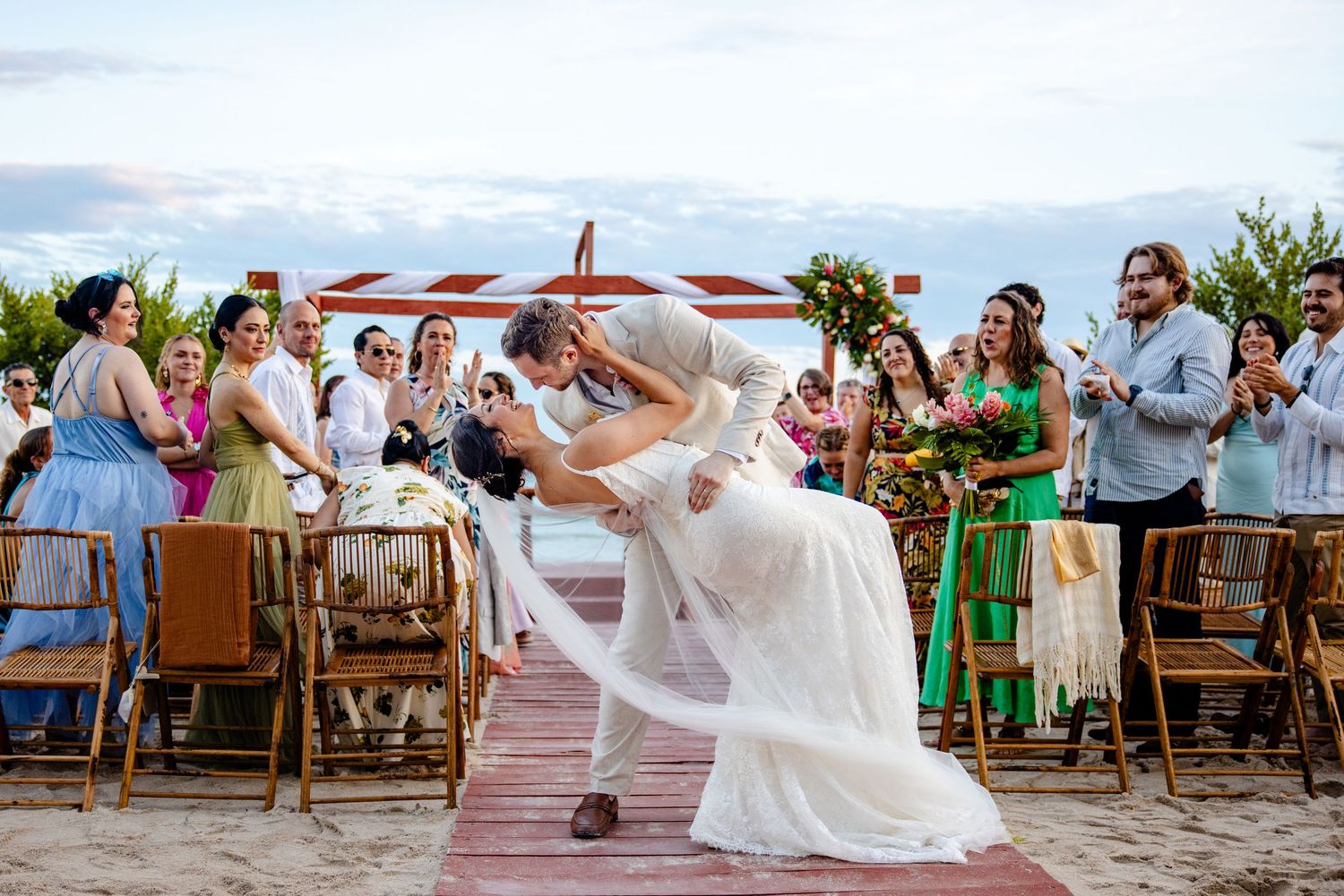 Couple shares romantic dip kiss at outdoor beach wedding ceremony with guests cheering around them.