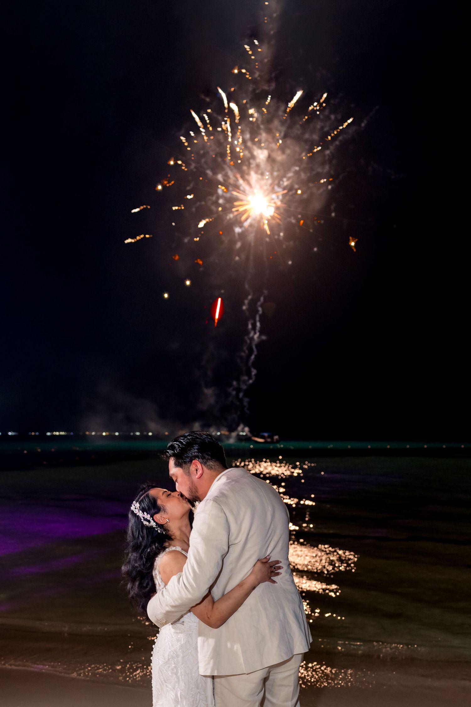Newlyweds share a romantic kiss on the beach with colorful fireworks exploding in the night sky above.