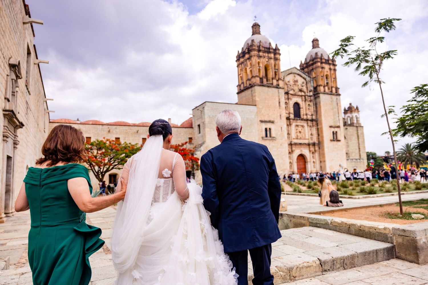 Bride in white gown walks with family toward ornate colonial church in Oaxaca, Mexico on her wedding day.