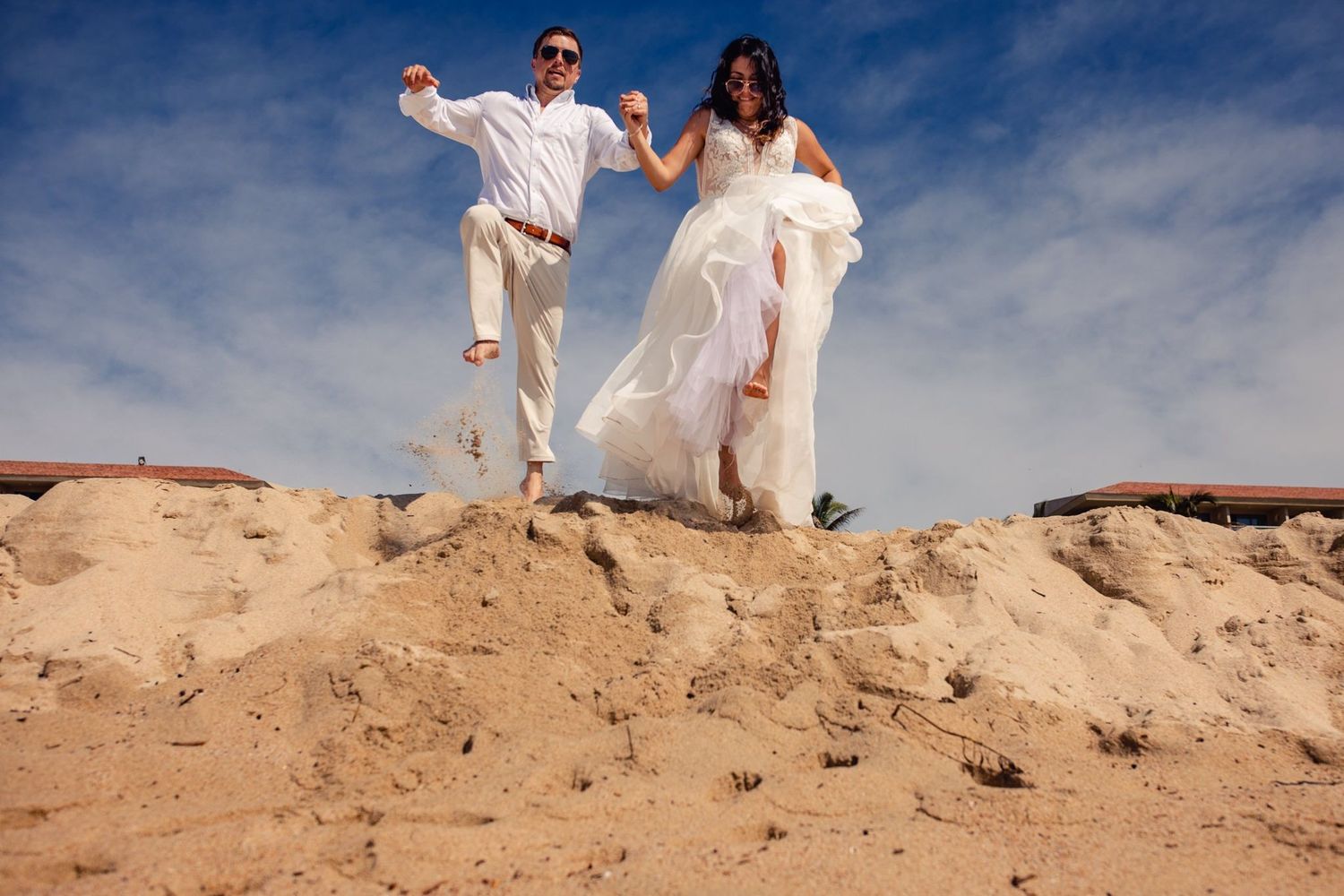 Bride and groom jumping joyfully on a sandy dune under a blue sky in wedding attire.