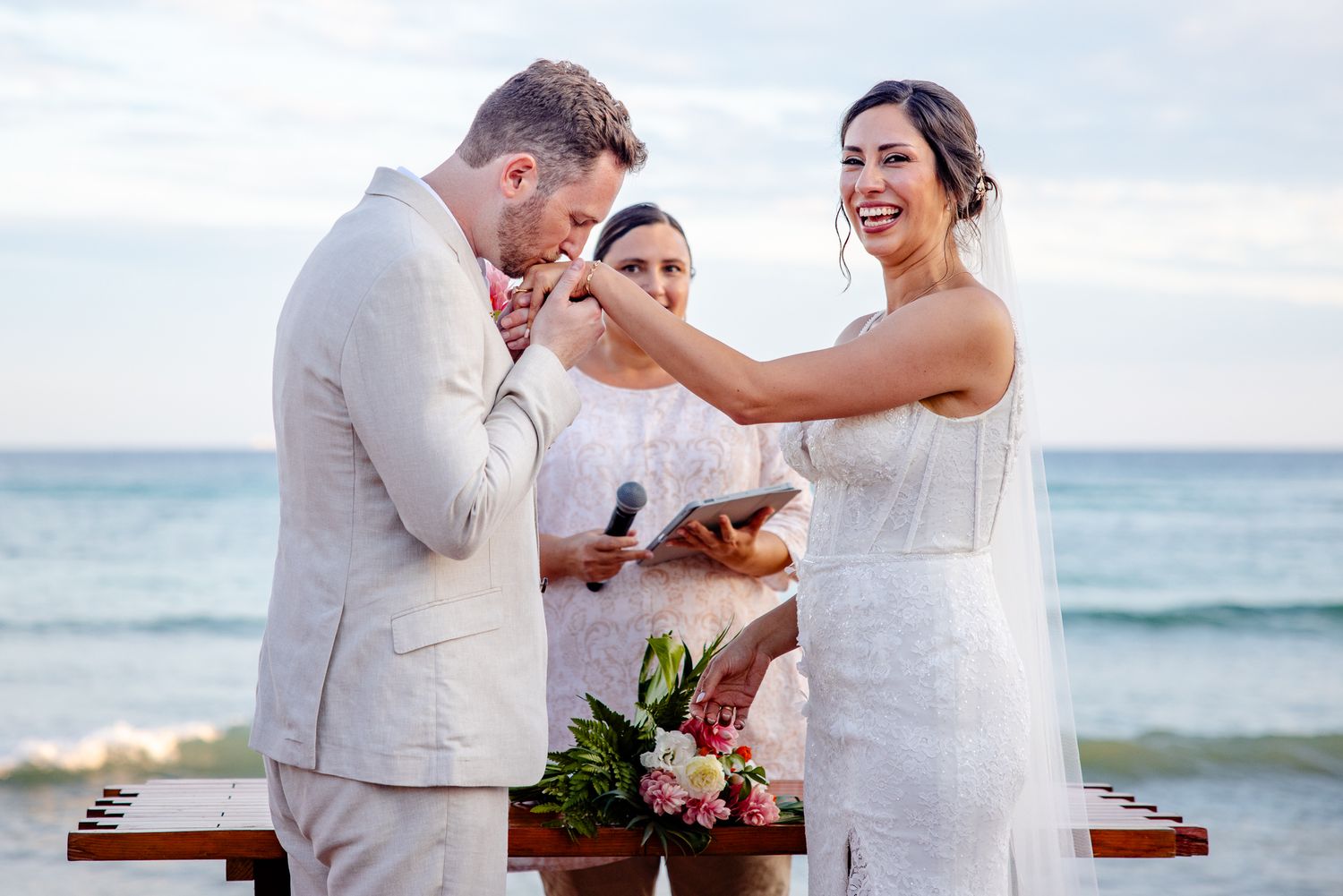 Bride smiles while placing ring on groom's finger during romantic beach wedding ceremony at sunset.