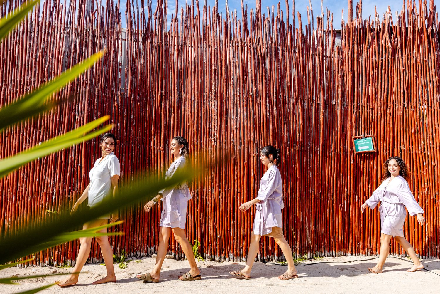Four women in white robes walk along a sandy path beside a rustic red bamboo wall at a luxury spa resort.