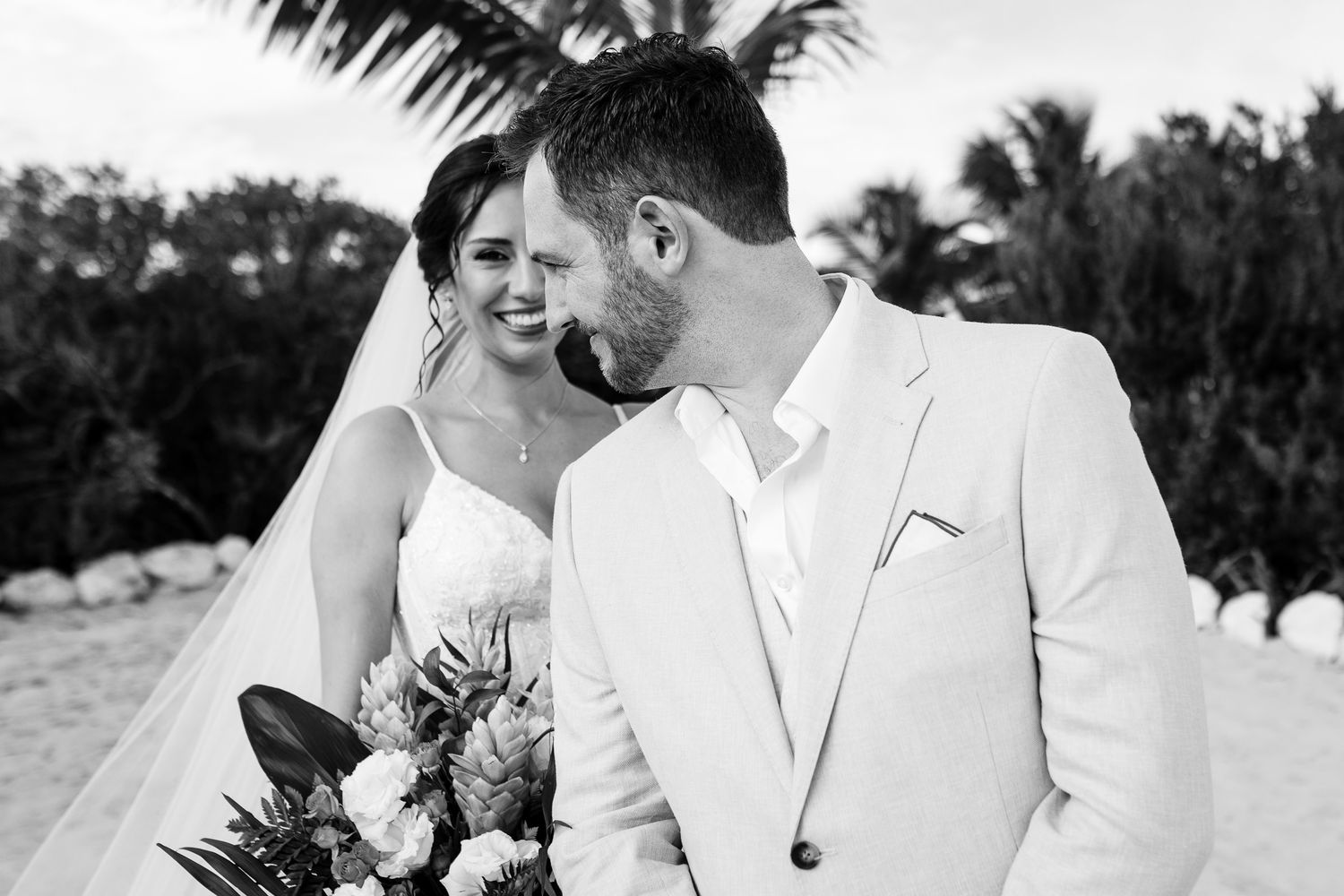 Black and white photo of a bride and groom sharing a romantic moment on a beach, bride holding a tropical bouquet.