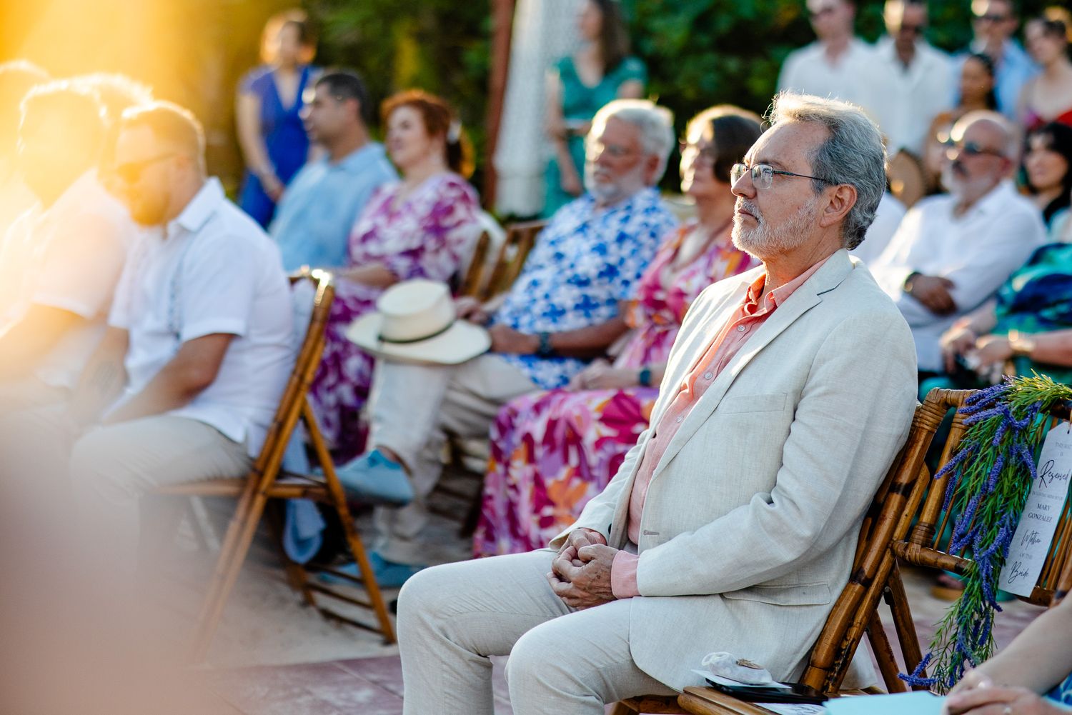 Man in beige suit sitting outdoors at a sunny garden event with guests seated in wooden chairs behind him.