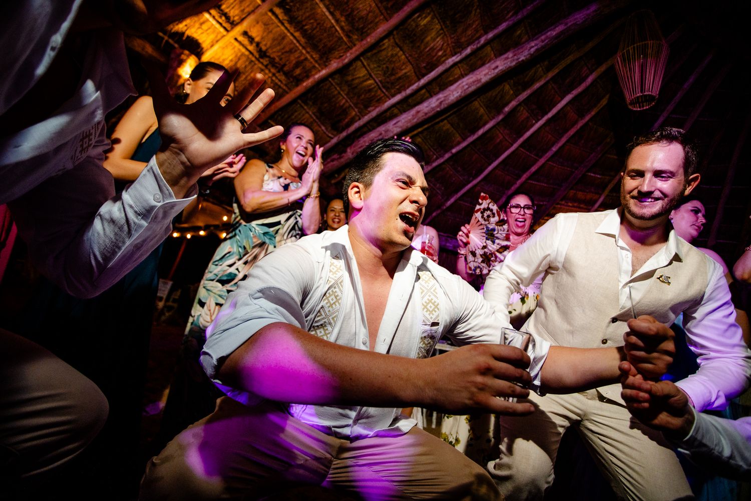 Energetic wedding reception dance floor with guests celebrating, man kneeling in white shirt dancing joyfully under warm lights.