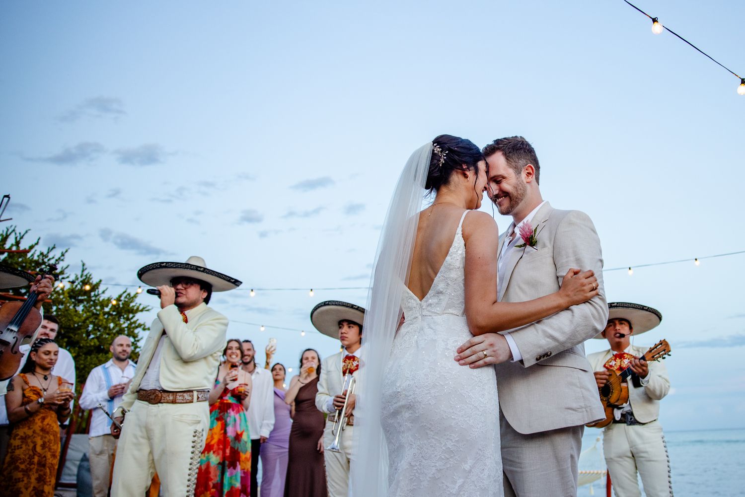 Newlyweds share first dance embrace surrounded by mariachi band at beachside wedding.