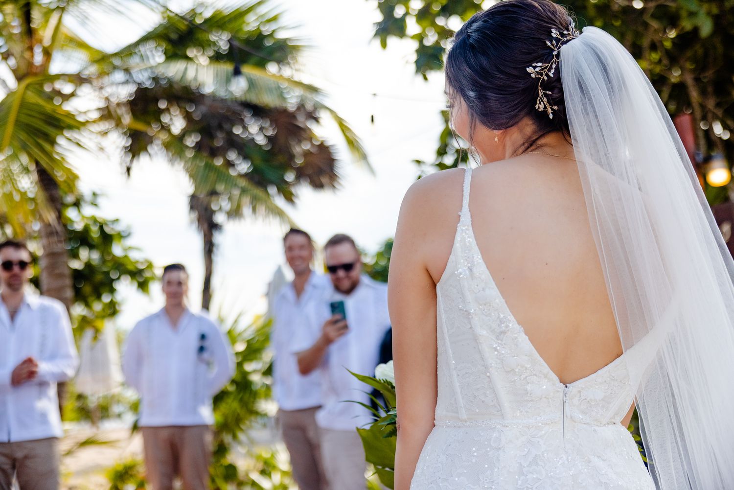 Bride in elegant backless wedding dress with veil stands before groomsmen at tropical outdoor ceremony.