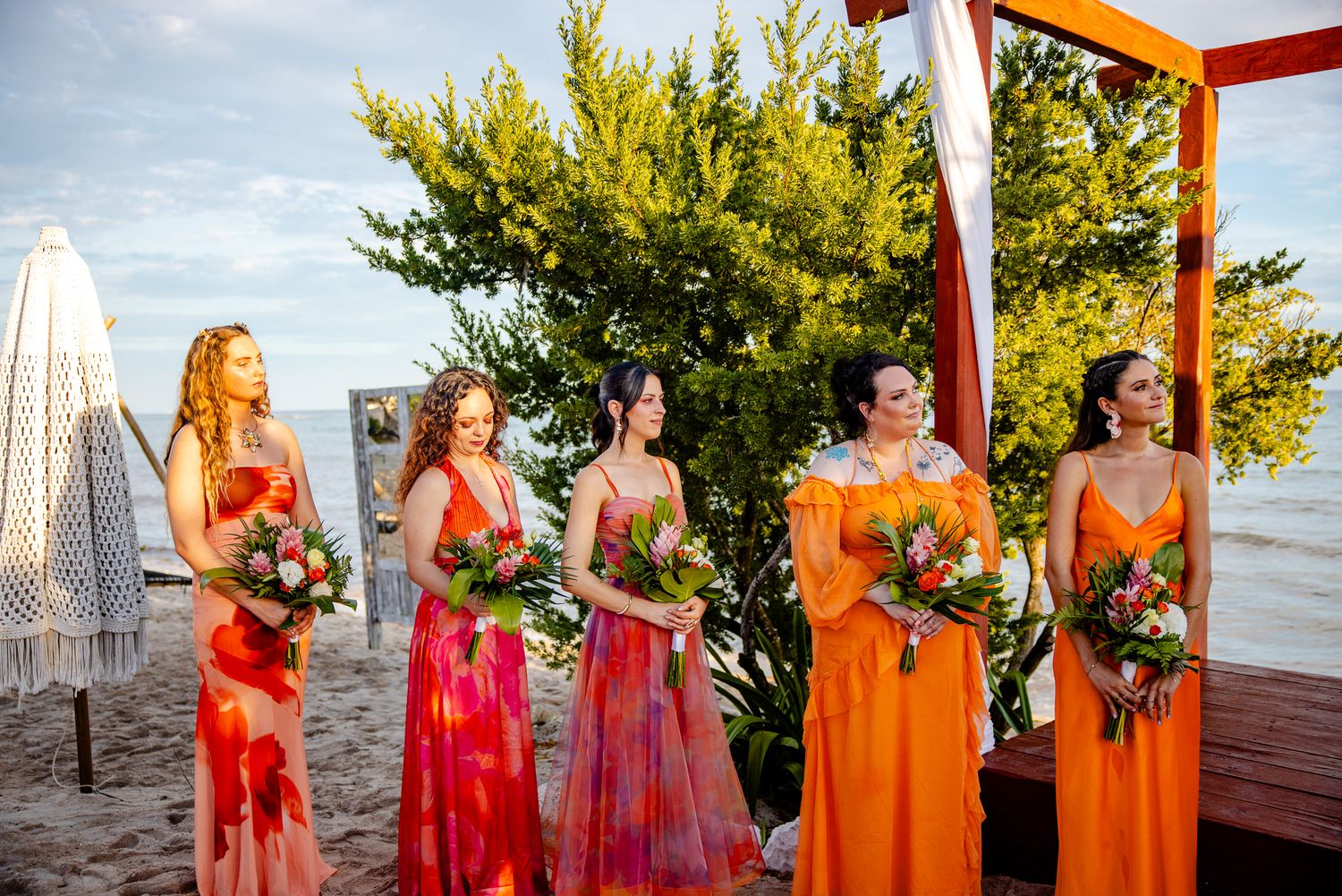 Five bridesmaids in orange and red floral gowns holding bouquets stand on a beach during an outdoor wedding ceremony.