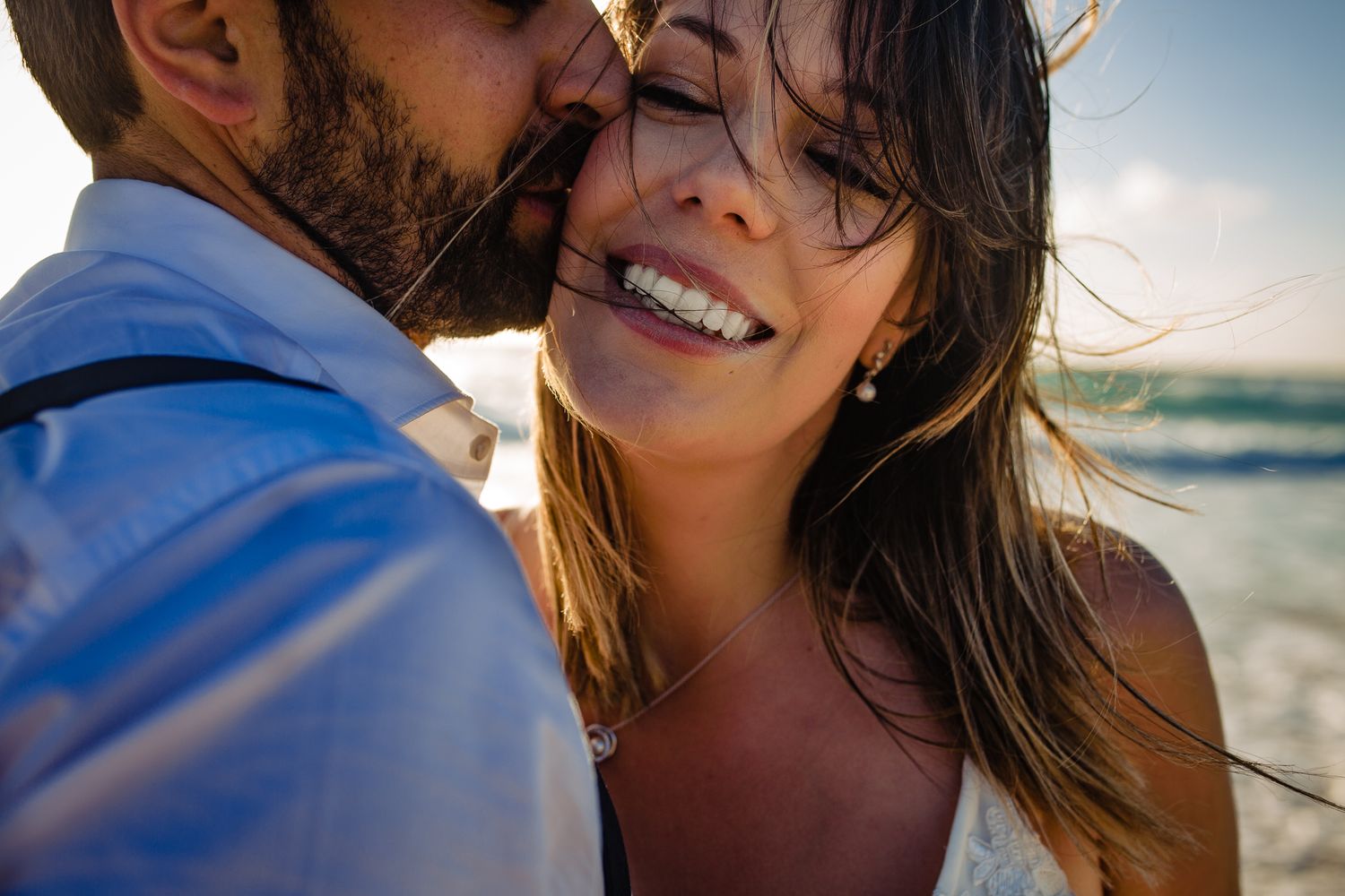 Couple sharing a romantic beach moment at sunset, groom kissing bride's cheek as she smiles joyfully.